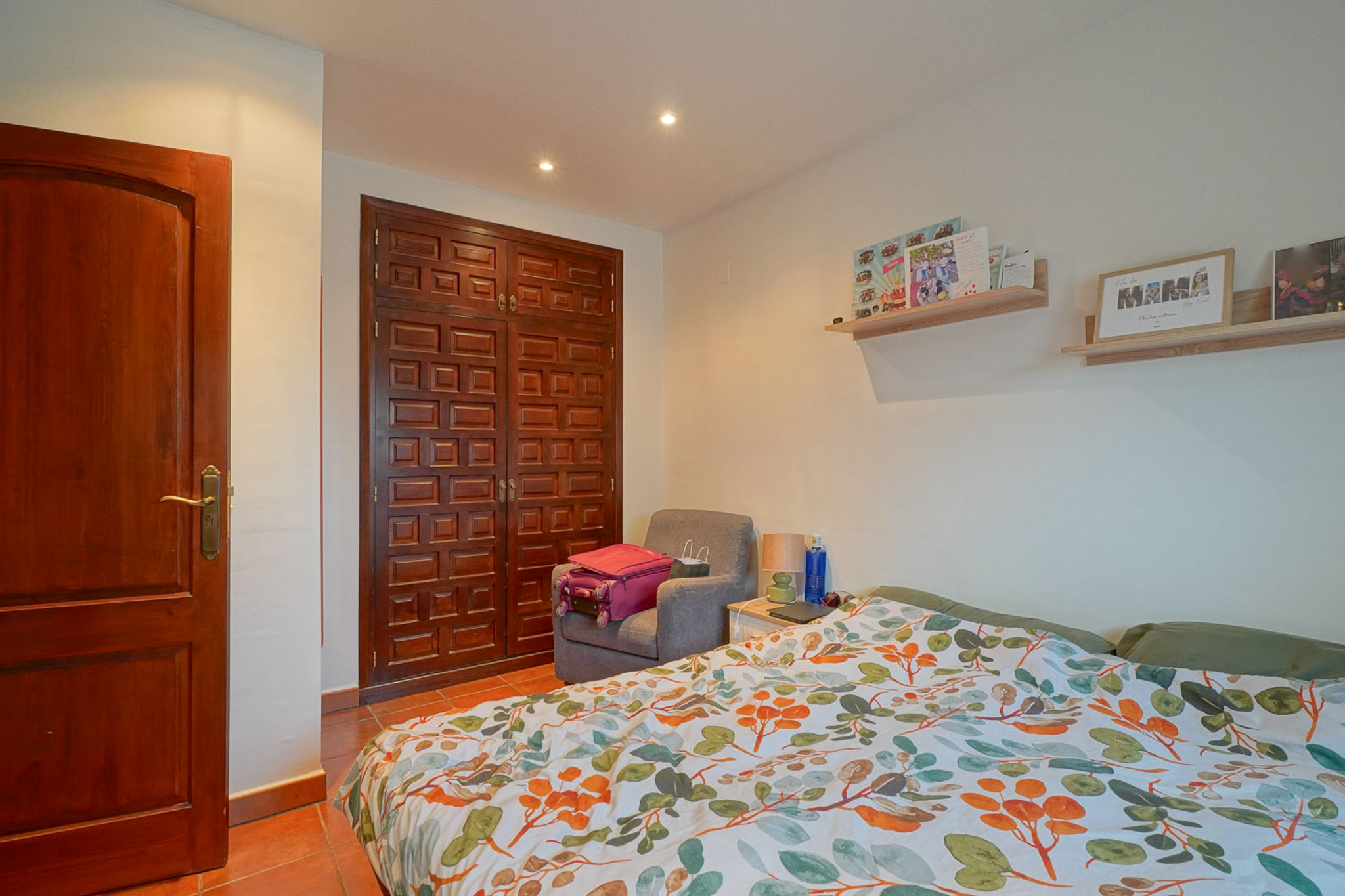 Schlafzimmer mit dunklem Holzschrank und -tür, Terrakottafliesenboden und Bett mit lebhaftem Blättermuster. Die Decke ist mit Einbauleuchten versehen.