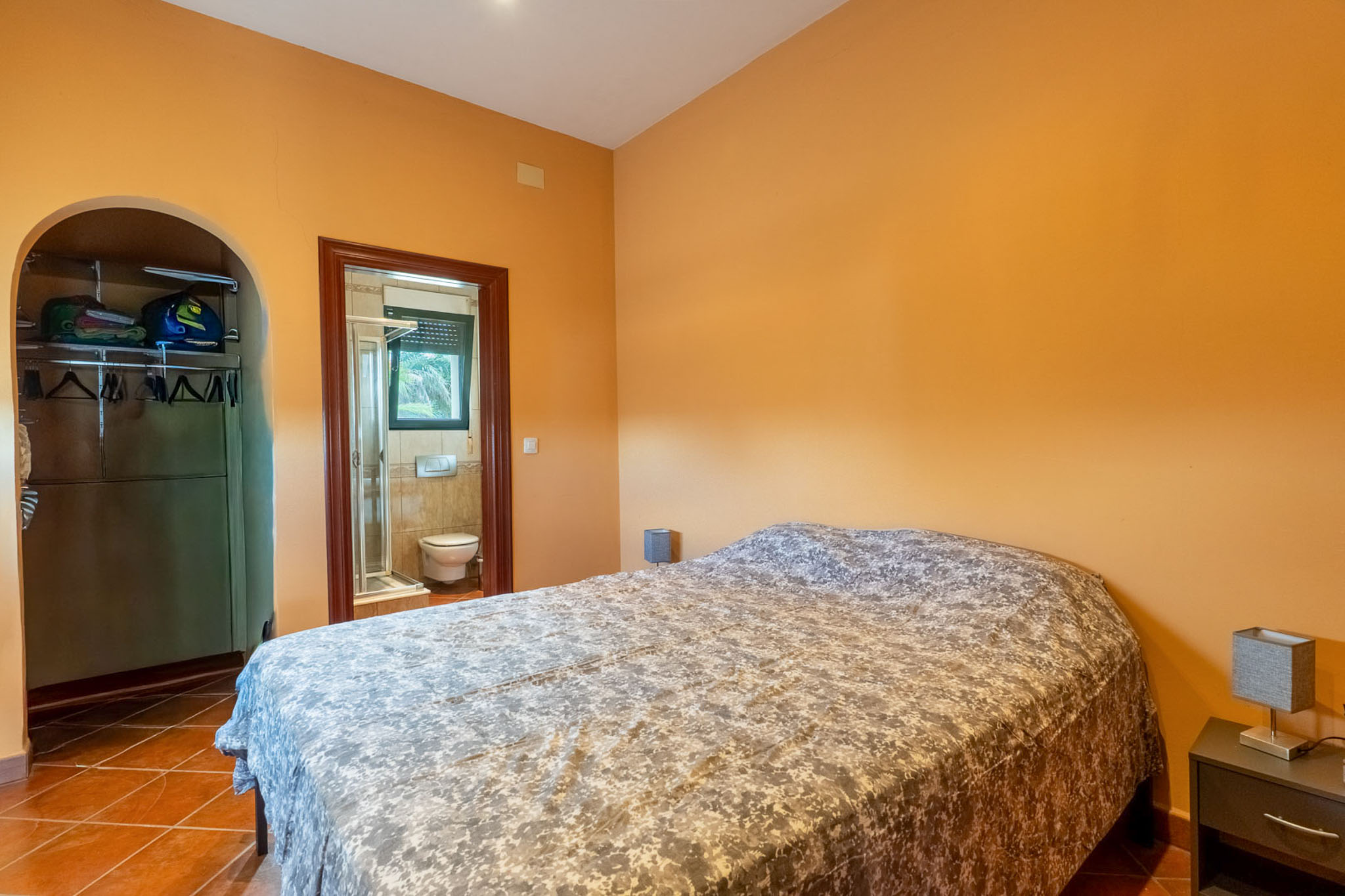Schlafzimmer mit orangefarbenen Wänden und Terrakottafliesen, ausgestattet mit einem Doppelbett. Ein offener Kleiderschrank und ein angrenzendes Badezimmer mit Dusche sind sichtbar.