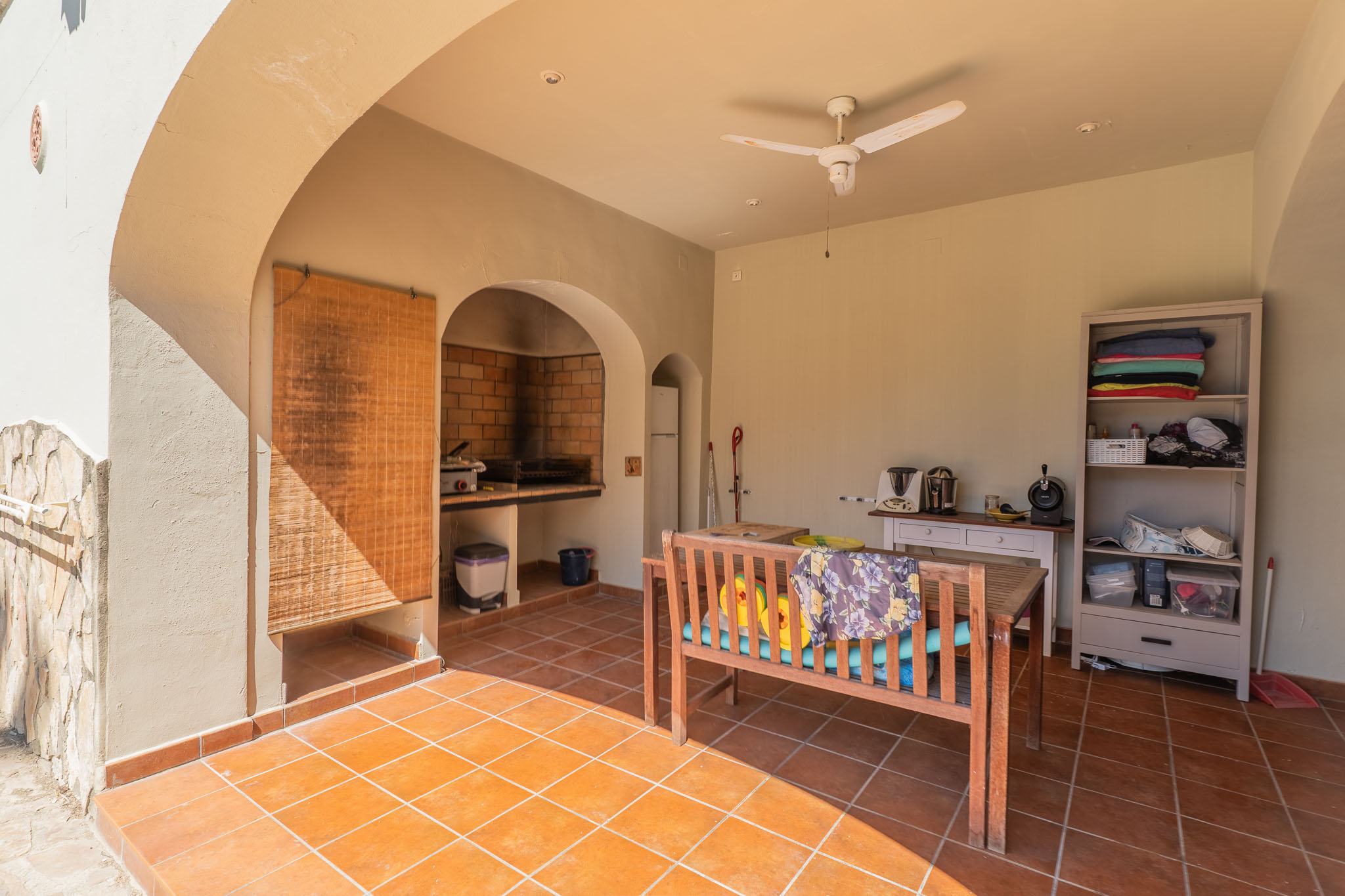 Überdachte Terrasse mit Terrakottafliesen und gemauertem Grill. Ausgestattet mit einer Holzsitzbank, Deckenventilator und Stauraum, ideal für Mahlzeiten im Freien.