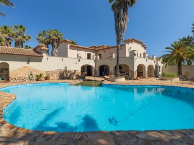 Außenansicht der mediterranen Villa mit unregelmäßig geformtem Privatpool, Natursteinumrandung und zahlreichen Palmen unter klarem blauem Himmel.