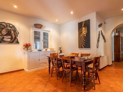 Esszimmer mit dunklem Holztisch und sechs Stühlen, Terrakotta-Fliesenboden und weißen Wänden mit Einbauleuchten. Ein weißer Schrank und zwei große Gemälde schmücken den Raum.