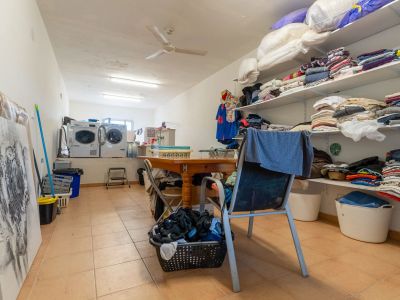 Funktioneller Hauswirtschaftsraum mit zwei Waschmaschinen, Trockner und offenen Regalen für Wäsche. Ein zentraler Holztisch bietet eine praktische Arbeitsfläche, ergänzt durch Fliesenboden und Deckenventilator.