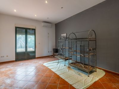 Geräumiger Raum mit Terrakotta-Fliesenboden, zweifarbigen Wänden, Einbauleuchten und Klimaanlage. Doppelt verglaste Tür mit Rollladen und große Metallvogelkäfige sind sichtbar.