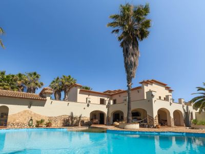 Großer privater Swimmingpool mit klarem Wasser, umgeben von Palmen und einer mediterranen Villa mit Arkaden unter blauem Himmel.