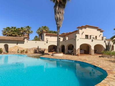 Großzügiger, unregelmäßig geformter Privatpool mit klarem blauen Wasser, umgeben von einer weitläufigen Natursteinterrasse. Die mediterrane Villa zeigt Bögen und Ziegeldächer inmitten von Palmen.