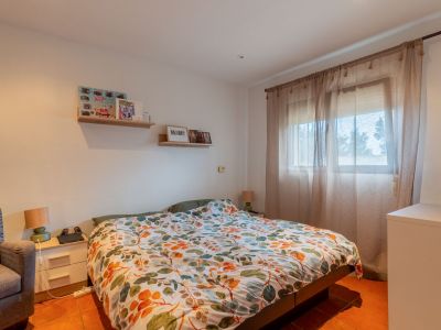 Helles Schlafzimmer mit Doppelbett, bunter Blätterbettdecke und Terrakottafliesenboden. Natürliches Licht fällt durch ein Fenster mit Gardinen, ergänzt durch Nachttisch und Wandregale.