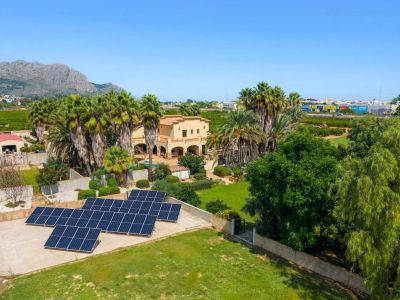 Luftaufnahme der traditionellen Villa, umgeben von Palmen und üppigen Gärten, mit einer prominenten Solaranlage und fernen Bergblicken unter klarem Himmel.