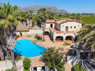 Luftaufnahme der Villa mit großem Freiformpool, weitläufigem gepflasterten Patio und Palmen, vor einer Bergkulisse und grünen Feldern.