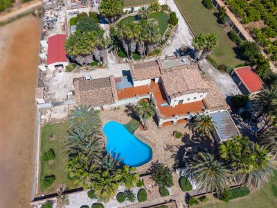 Luftaufnahme des Anwesens mit unregelmäßig geformtem blauem Pool, umgeben von Palmen und Gärten. Mehrere Gebäude mit Ziegeldächern und ein herzförmiger Teich sind sichtbar.