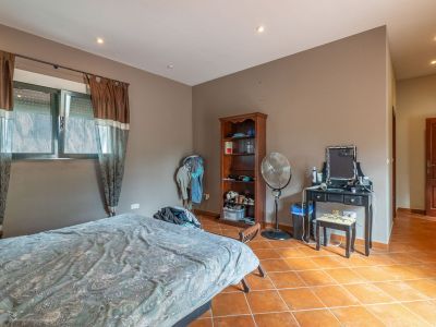 Schlafzimmer mit Bett, Fenstern mit braunen Gardinen und Rollos, sowie Terrakotta-Fliesenboden. Ausgestattet mit schwarzem Schminktisch, Holzregal und Standventilator.