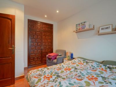 Schlafzimmer mit dunklem Holzschrank und -tür, Terrakottafliesenboden und Bett mit lebhaftem Blättermuster. Die Decke ist mit Einbauleuchten versehen.