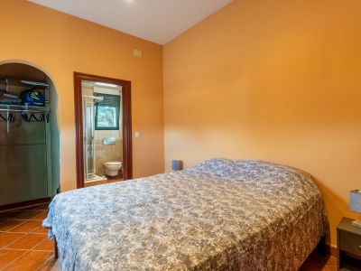 Schlafzimmer mit orangefarbenen Wänden und Terrakottafliesen, ausgestattet mit einem Doppelbett. Ein offener Kleiderschrank und ein angrenzendes Badezimmer mit Dusche sind sichtbar.