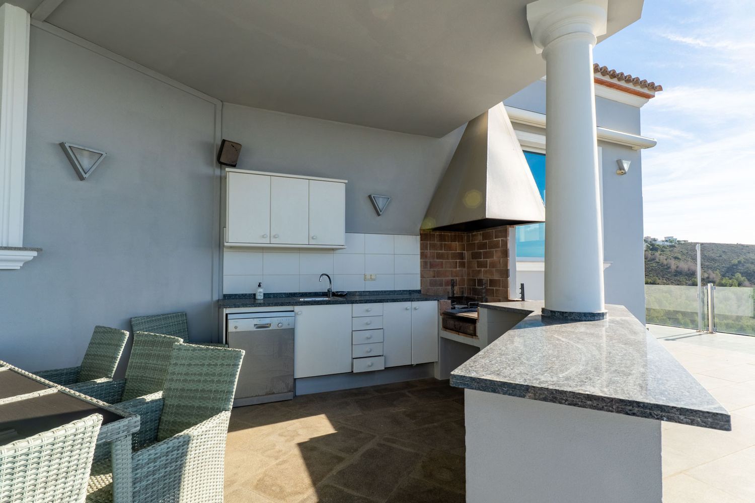 Ausgestattete Außenküche mit gemauertem Grill, Granitarbeitsplatten, Spüle und Geschirrspüler, neben einem Esstisch auf einer überdachten Terrasse mit Hügelblick.