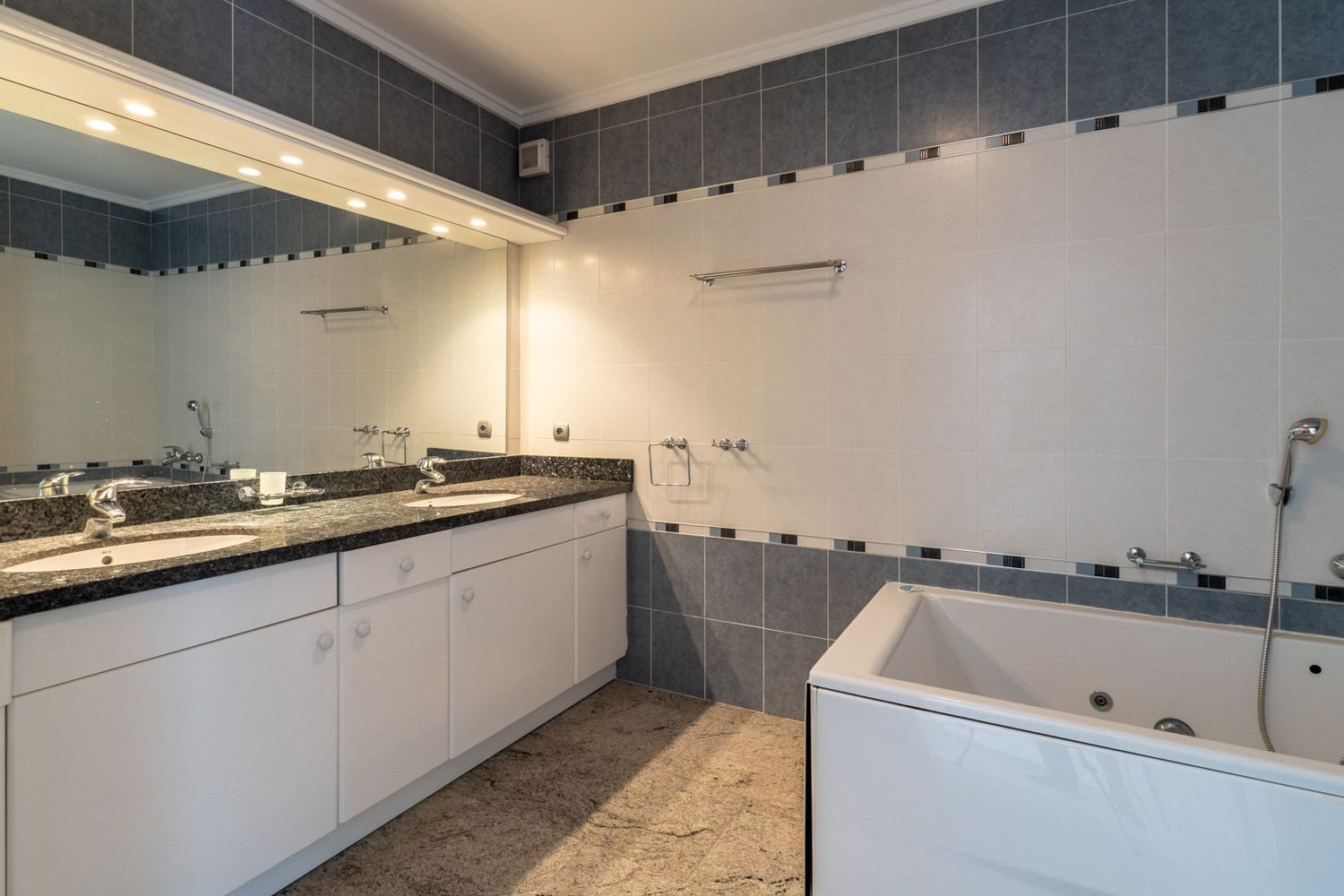 Badezimmer mit Doppelwaschtisch aus dunklem Granit auf weißen Schränken, integrierter Badewanne und großem beleuchtetem Spiegel. Wände sind grau-weiß gefliest.