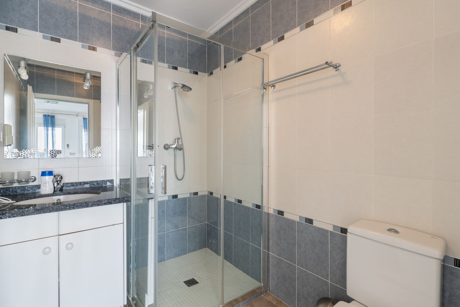 Badezimmer mit dunkler Granit-Waschtischplatte, dekorativem Spiegel und Glasduschkabine. Die Wände sind mit hellbeigen und blau-grauen Fliesen sowie einer Mosaikbordüre versehen.
