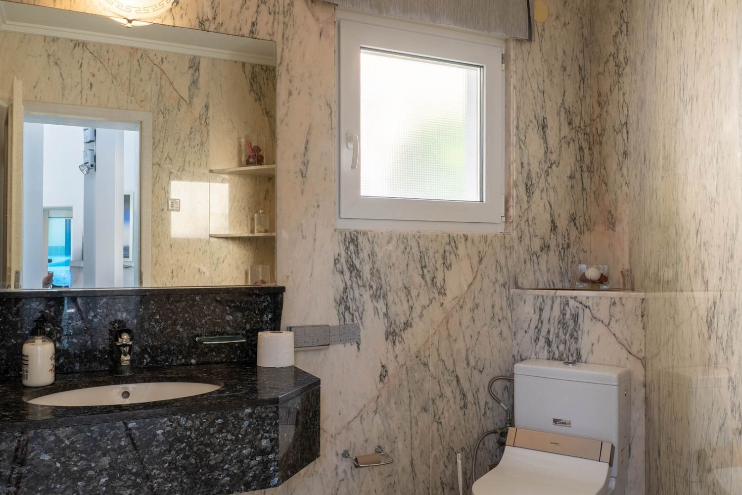 Badezimmer mit hellen, geäderten Marmorwänden, einem dunklen Granitwaschtisch mit integriertem Waschbecken und einem großen Spiegel. Ein Milchglasfenster und eine Toilette mit Bidetfunktion sind vorhanden.