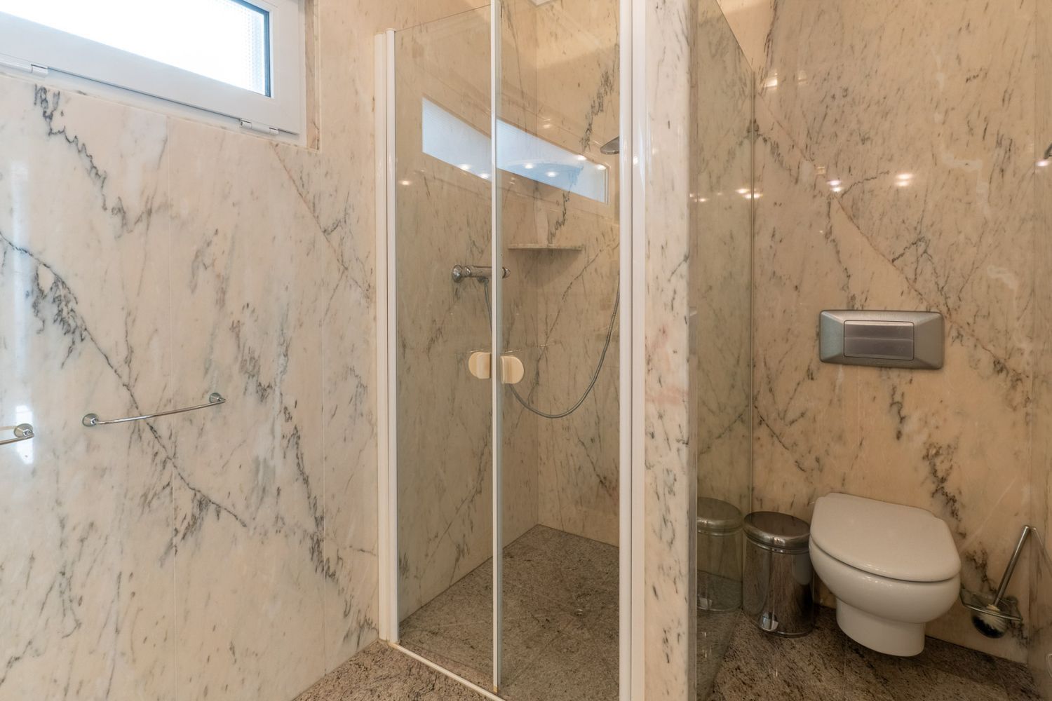 Badezimmer mit Wänden aus geädertem Marmor und Granitboden. Es verfügt über eine Glasdusche mit integrierter Ablage und ein wandhängendes WC, beleuchtet durch ein Oberlichtfenster.