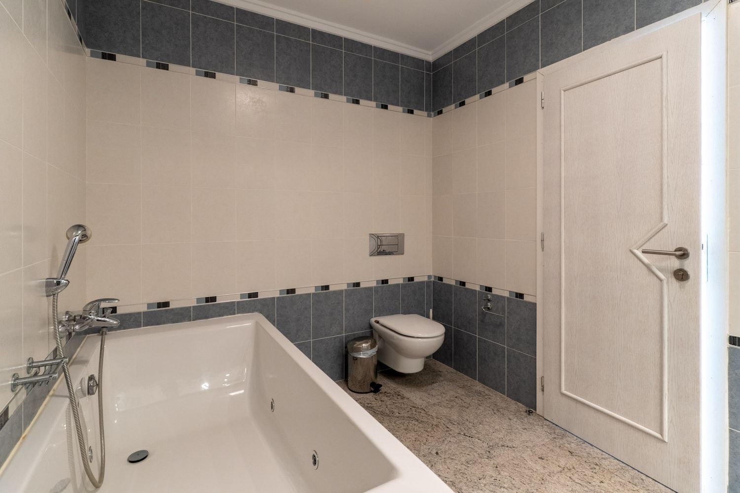 Badezimmer mit weißer Badewanne und integrierter Dusche. Beige und grau geflieste Wände mit dekorativer Bordüre. Heller Granitboden, wandhängende Toilette und eine Tür mit geometrischem Design.
