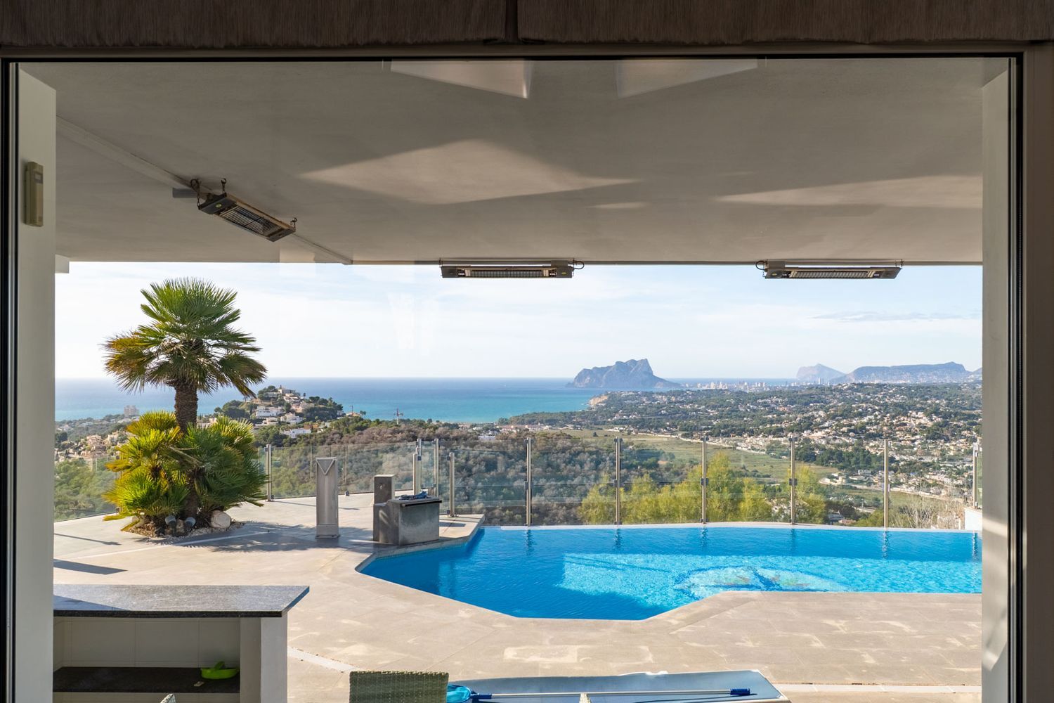 Blick von innen auf die Terrasse mit Pool, Palmen und einem weiten Panorama des Meeres, der Küste von Moraira und des Peñón de Ifach. Deckenheizstrahler unter dem Vordach.