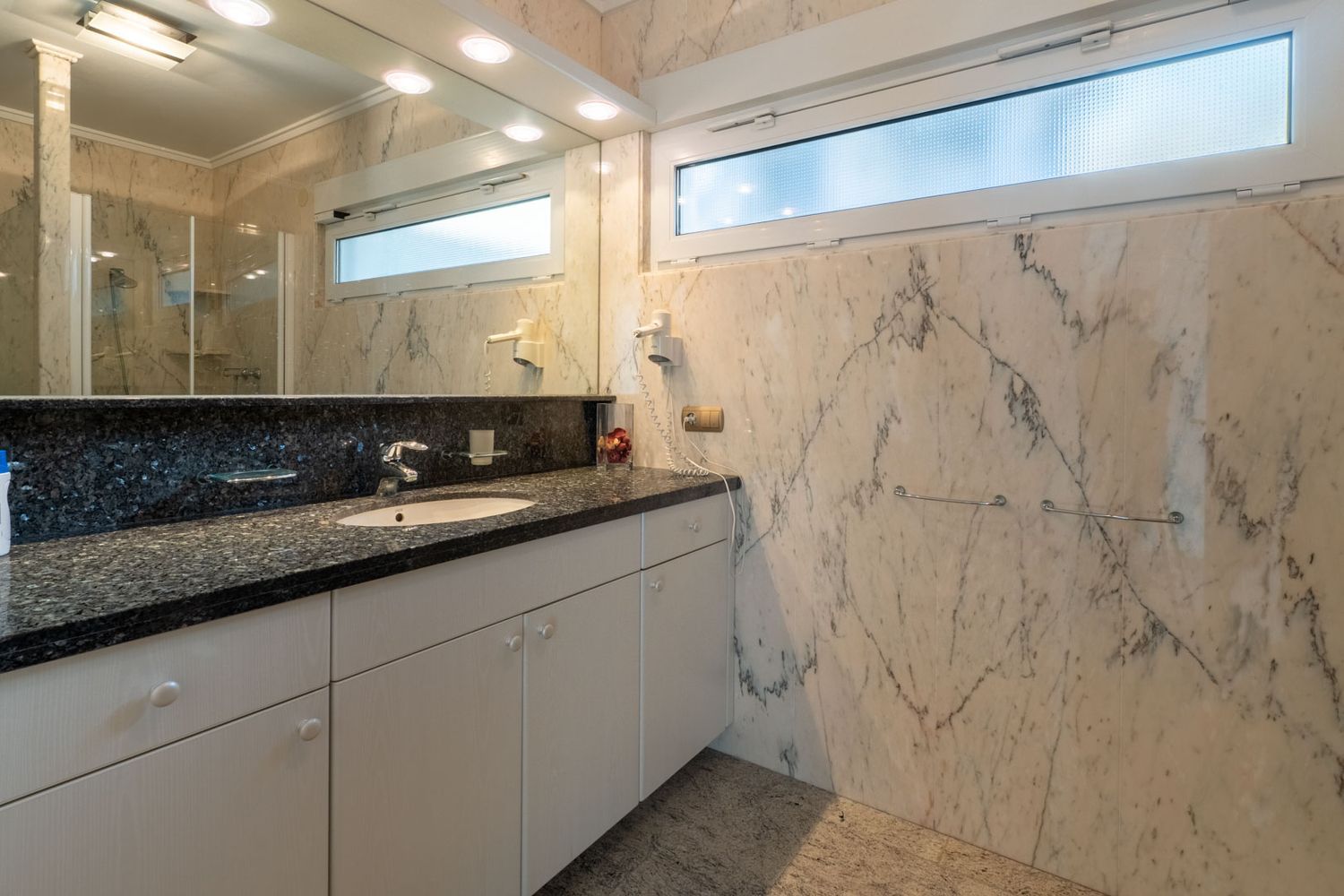 Elegantes Badezimmer mit Wänden und Boden aus geädertem Marmor. Der Waschtisch verfügt über eine dunkle Granitarbeitsplatte und ein integriertes Waschbecken, ergänzt durch ein mattiertes Oberlicht.