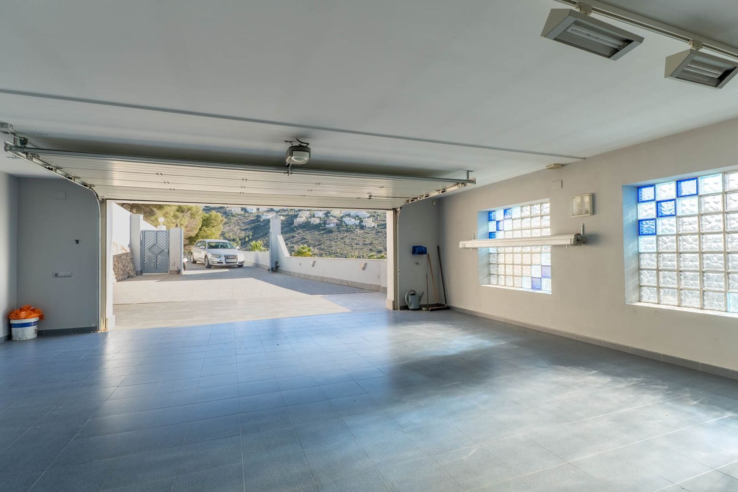 Geräumige Garage mit hellblauem Fliesenboden und Glasbausteinfenstern mit blauen Akzenten, das Tor öffnet sich zur Auffahrt und den entfernten Hügeln.