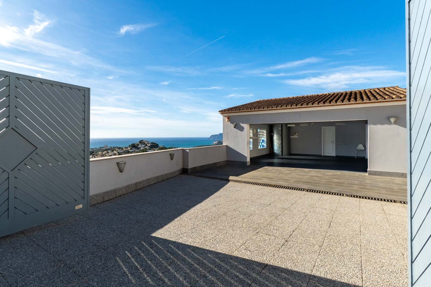 Geräumiger Außenbereich mit Fliesenpflaster und Zugang zur überdachten Garage. Panoramablicke auf das Mittelmeer und die Küste, eingerahmt von einem modernen grauen Tor und einer niedrigen Mauer mit Beleuchtung.