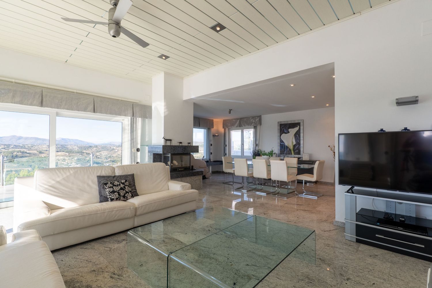 Geräumiger Wohn- und Essbereich mit poliertem Marmorboden, weißem Sofa und dunklem Steinkamin. Große Fenster bieten Panoramablicke auf die Berge und die Landschaft.
