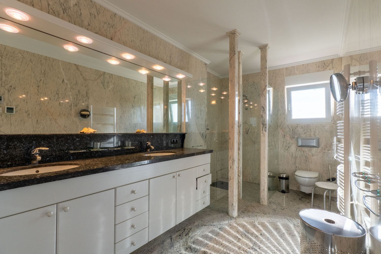 Geräumiges Badezimmer mit Doppelwaschtisch, dunkler Granitplatte und weißen Schränken. Begehbare Dusche mit Glaswänden und Marmorsäulen, passend zu den hellen Marmorwänden und dem Boden.