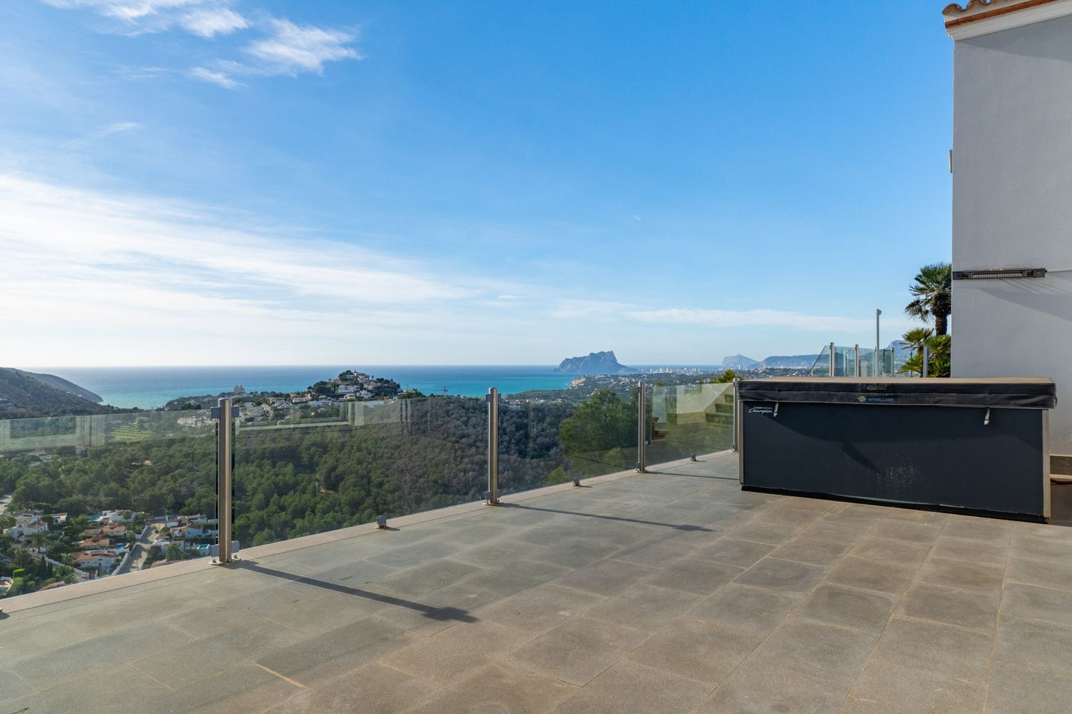 Große Außenterrasse mit grauen Fliesen und überdachtem Whirlpool, umrahmt von einer Glasbalustrade mit Panoramablick auf das Meer und den Peñón de Ifach.