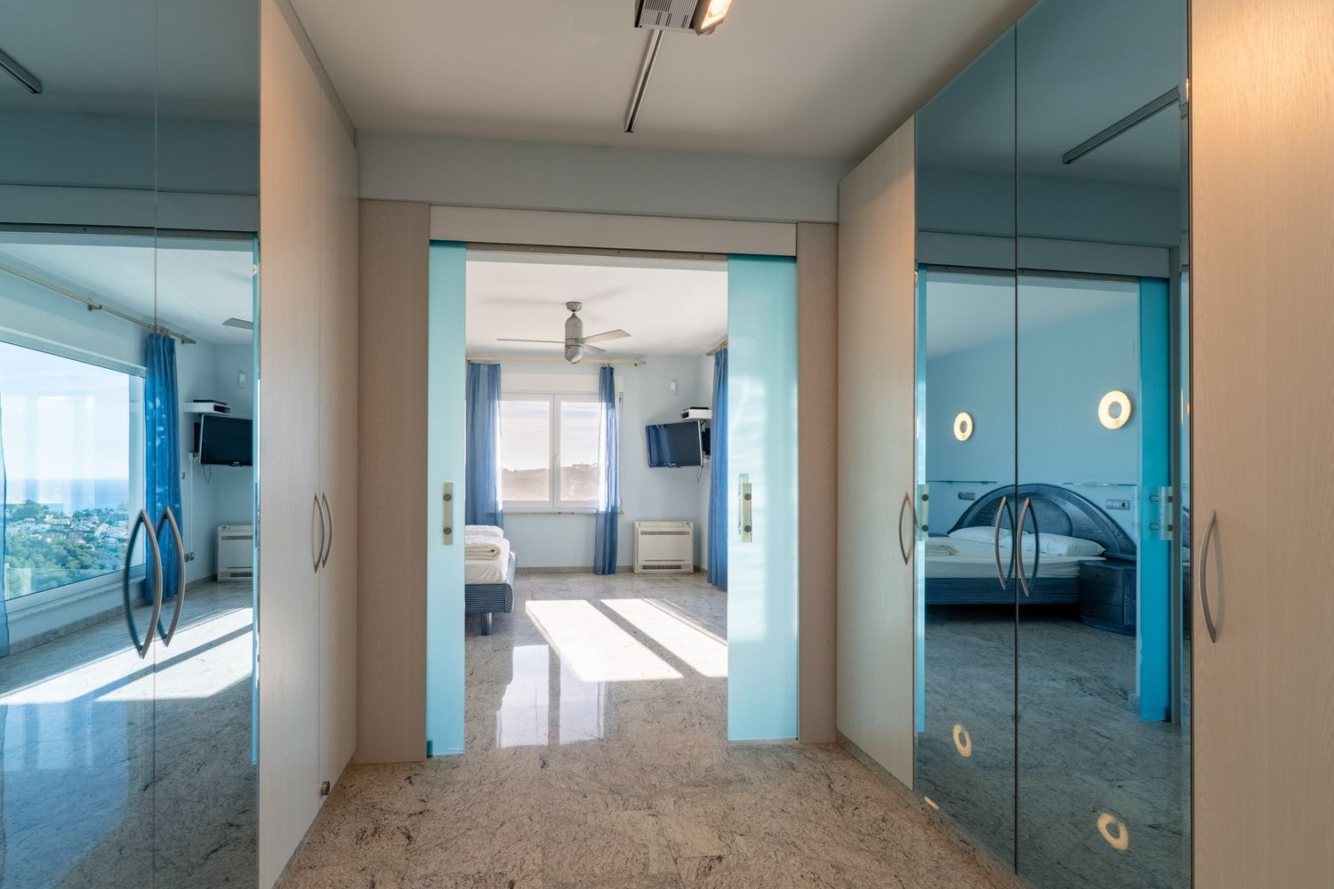 Heller Flur mit Granitboden und hellen Holzschränken. Glasschiebetüren rahmen ein Schlafzimmer mit Meerblick ein, während Spiegeltüren ein weiteres blaues Schlafzimmer reflektieren.