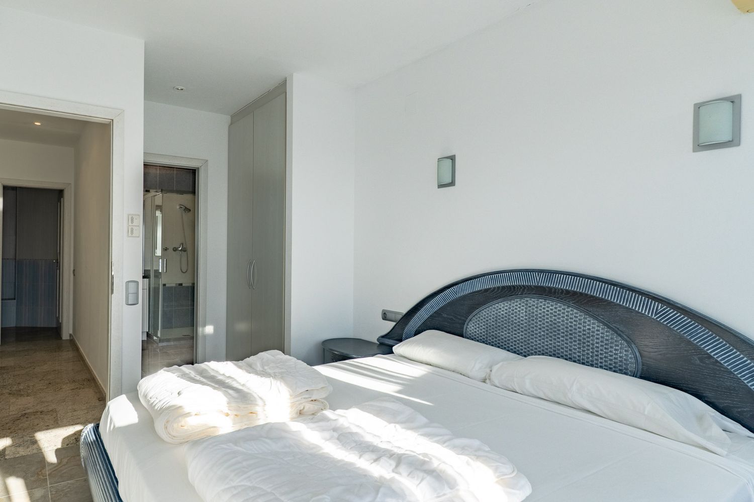 Helles Schlafzimmer mit Doppelbett und dunklem Holzkopfteil mit geflochtener Einlage. Es verfügt über einen Einbauschrank und direkten Zugang zu einem Bad mit Glasdusche.