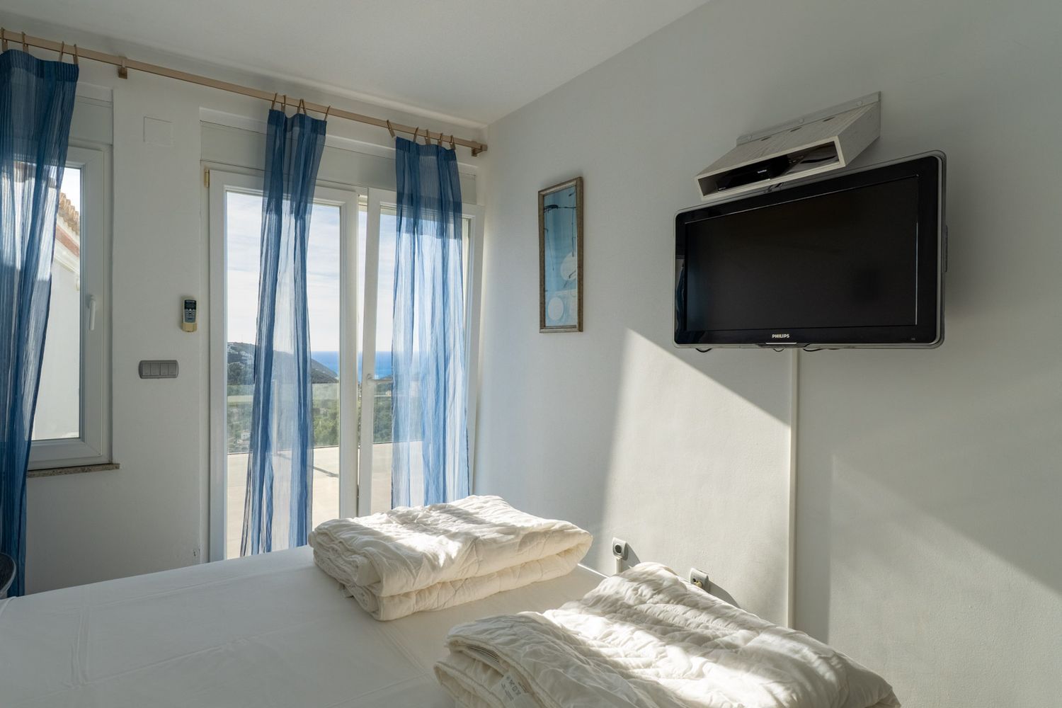 Helles Schlafzimmer mit Einzelbetten und direktem Zugang zur Terrasse. Glasschiebetüren mit blauen Vorhängen bieten Meerblick. Ein wandmontierter Fernseher ist vorhanden.
