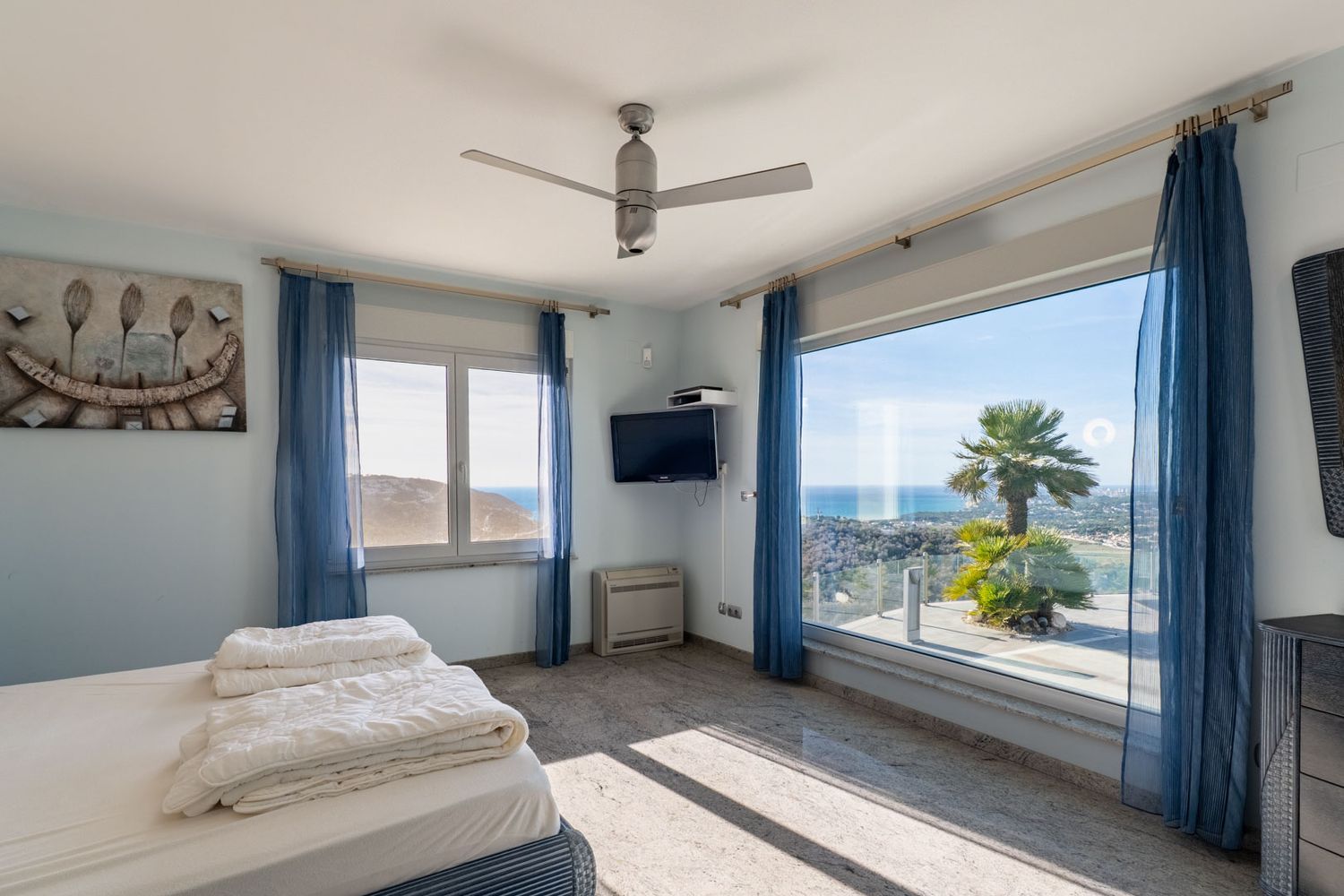 Helles Schlafzimmer mit großem Panoramafenster, direktem Meerblick und Palme. Heller Granitfliesenboden und blaue transparente Vorhänge.