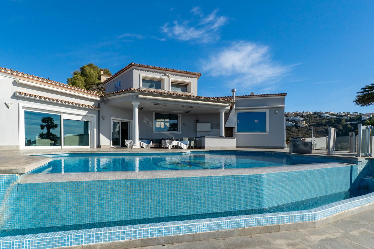 Infinity-Pool mit blauen Mosaikfliesen und überdachte Terrasse mit Außenküche, direkt an der Villenfassade.
