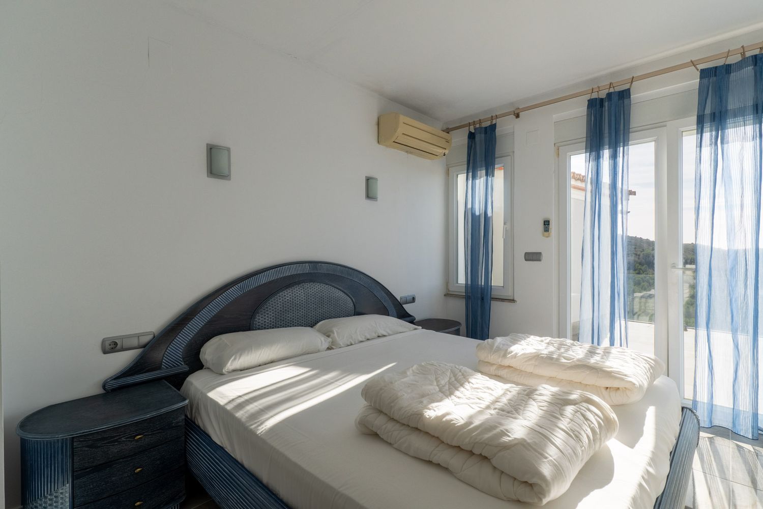 Schlafzimmer mit blau-dunklem Doppelbett und passenden Nachttischen. Glastüren mit blauen Vorhängen führen zur Terrasse, ergänzt durch eine Klimaanlage.