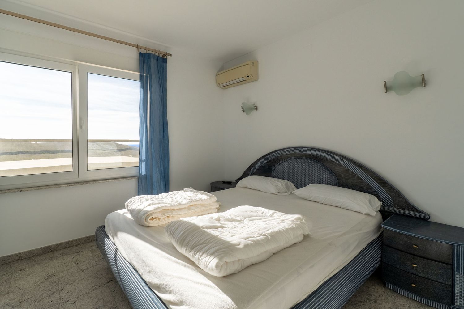 Schlafzimmer mit blau gemustertem Bett und geflochtenem Kopfteil, hellen Fliesenboden und Außenblick durch das Fenster. Eine Klimaanlage und passender Nachttisch sind vorhanden.
