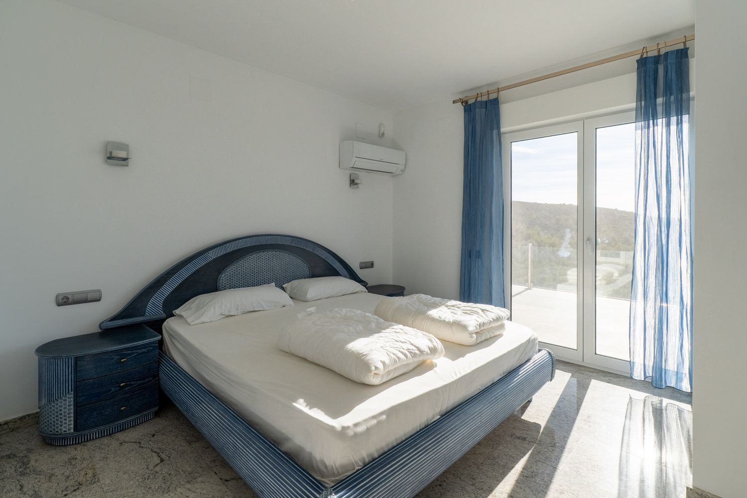 Schlafzimmer mit blauem Designbett und passenden Nachttischen, hellem Boden und direktem Terrassenzugang durch Glastür, mit Hügelaussicht und Tageslicht.