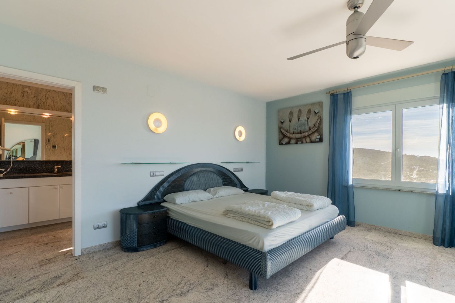 Schlafzimmer mit einem blau getönten Designerbett, passenden Nachttischen und hellem Marmorboden. Das Fenster bietet Hügelblick, ergänzt durch blaue Vorhänge und ein angrenzendes Badezimmer.