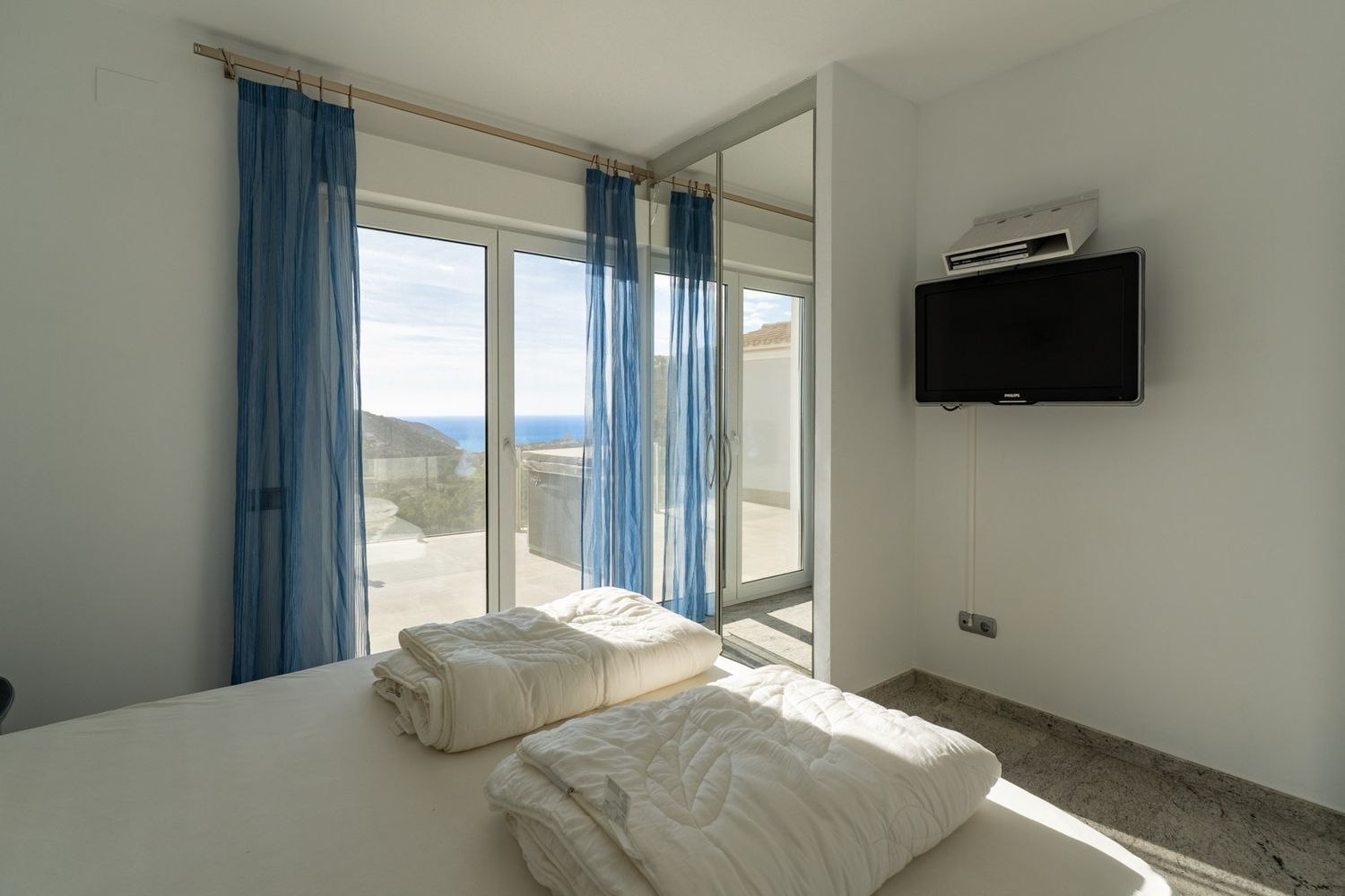 Schlafzimmer mit weiß bezogenem Bett, Wandfernseher und großen Glasschiebetüren mit blauen Gardinen, die Zugang zu einer Terrasse mit Panoramameerblick bieten.