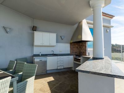 Ausgestattete Außenküche mit gemauertem Grill, Granitarbeitsplatten, Spüle und Geschirrspüler, neben einem Esstisch auf einer überdachten Terrasse mit Hügelblick.