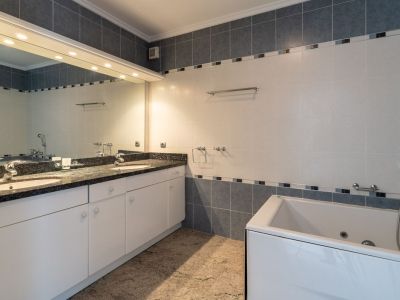 Badezimmer mit Doppelwaschtisch aus dunklem Granit auf weißen Schränken, integrierter Badewanne und großem beleuchtetem Spiegel. Wände sind grau-weiß gefliest.