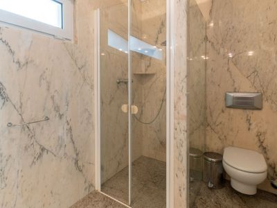 Badezimmer mit Wänden aus geädertem Marmor und Granitboden. Es verfügt über eine Glasdusche mit integrierter Ablage und ein wandhängendes WC, beleuchtet durch ein Oberlichtfenster.