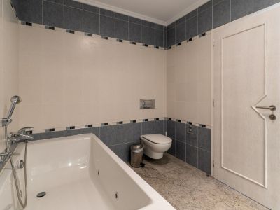 Badezimmer mit weißer Badewanne und integrierter Dusche. Beige und grau geflieste Wände mit dekorativer Bordüre. Heller Granitboden, wandhängende Toilette und eine Tür mit geometrischem Design.