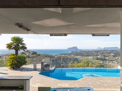 Blick von innen auf die Terrasse mit Pool, Palmen und einem weiten Panorama des Meeres, der Küste von Moraira und des Peñón de Ifach. Deckenheizstrahler unter dem Vordach.