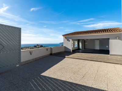 Geräumiger Außenbereich mit Fliesenpflaster und Zugang zur überdachten Garage. Panoramablicke auf das Mittelmeer und die Küste, eingerahmt von einem modernen grauen Tor und einer niedrigen Mauer mit Beleuchtung.