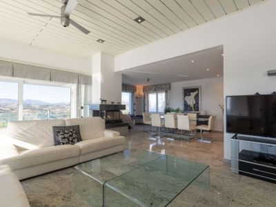 Geräumiger Wohn- und Essbereich mit poliertem Marmorboden, weißem Sofa und dunklem Steinkamin. Große Fenster bieten Panoramablicke auf die Berge und die Landschaft.