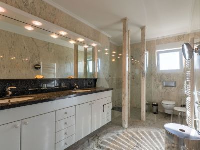 Geräumiges Badezimmer mit Doppelwaschtisch, dunkler Granitplatte und weißen Schränken. Begehbare Dusche mit Glaswänden und Marmorsäulen, passend zu den hellen Marmorwänden und dem Boden.
