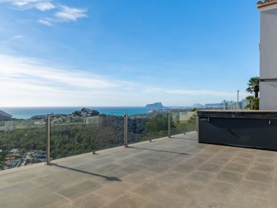 Große Außenterrasse mit grauen Fliesen und überdachtem Whirlpool, umrahmt von einer Glasbalustrade mit Panoramablick auf das Meer und den Peñón de Ifach.