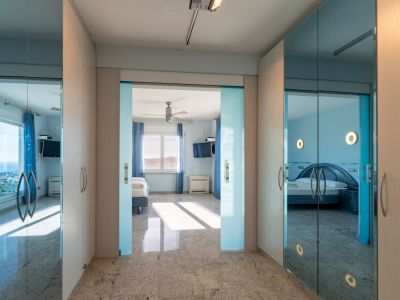 Heller Flur mit Granitboden und hellen Holzschränken. Glasschiebetüren rahmen ein Schlafzimmer mit Meerblick ein, während Spiegeltüren ein weiteres blaues Schlafzimmer reflektieren.