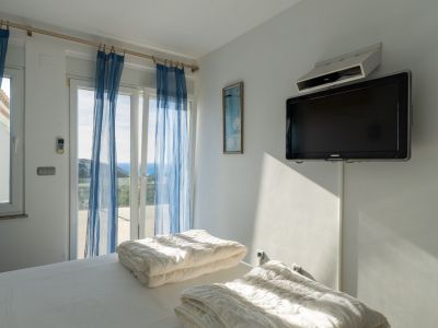 Helles Schlafzimmer mit Einzelbetten und direktem Zugang zur Terrasse. Glasschiebetüren mit blauen Vorhängen bieten Meerblick. Ein wandmontierter Fernseher ist vorhanden.